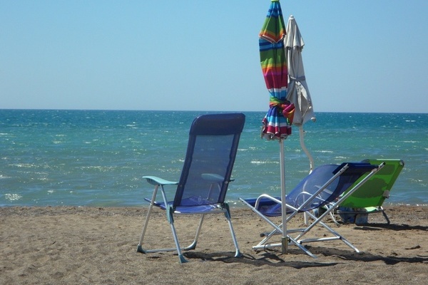 Zwei blaue Stühle und eine grüne Sonnenliege mit zwei zusammengefalteten Sonnenschirmen stehen am Strand