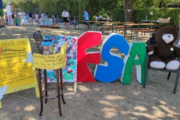 Plakat Abschluss Ferienspielaktion, Spendenkässle, aufgestellte Buchstaben FSA und Teddybär auf einem Stuhl