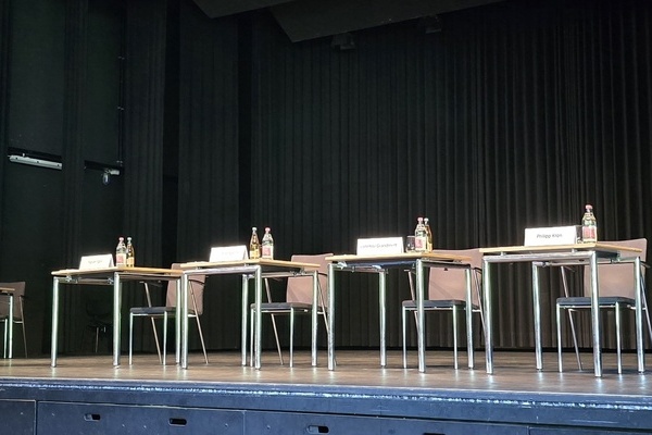 Auf der Bühne stehen ein Rednerpult und vier Tische mit Namensschildern  mit Stühlen für die vier Bewerber