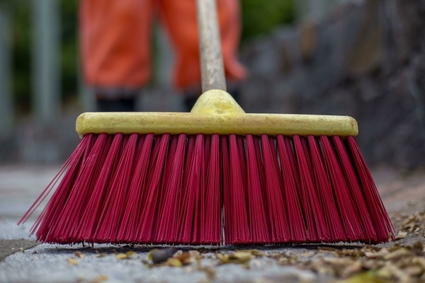 Besen mit roten Borsten. Im Hintergrund verschwommen zu erahnen eine Person in orangefarbener Arbeitsbekleidung. Foto: Mauricio Monteiro Fran&ccedil;a von Pixabay